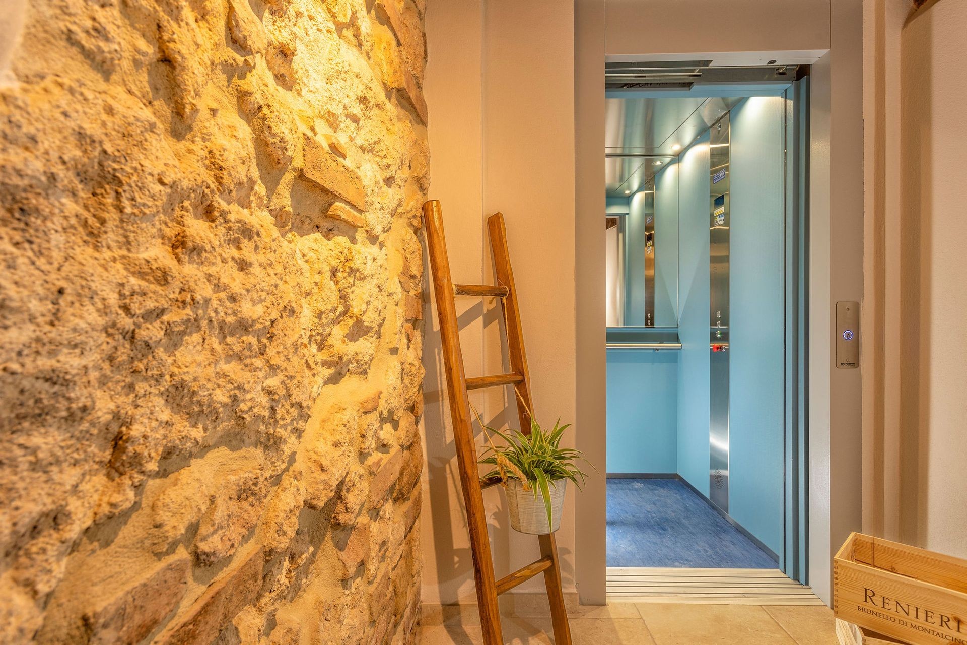 Wooden ladder with a plant against a stone wall beside an open modern elevator.